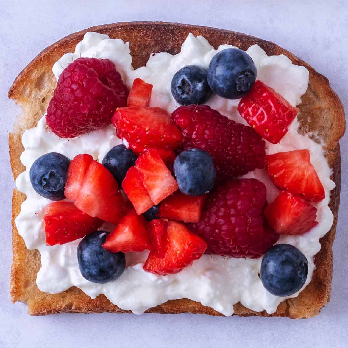Sliced strawberries, raspberries and blueberries on top of cottage cheese on toast.