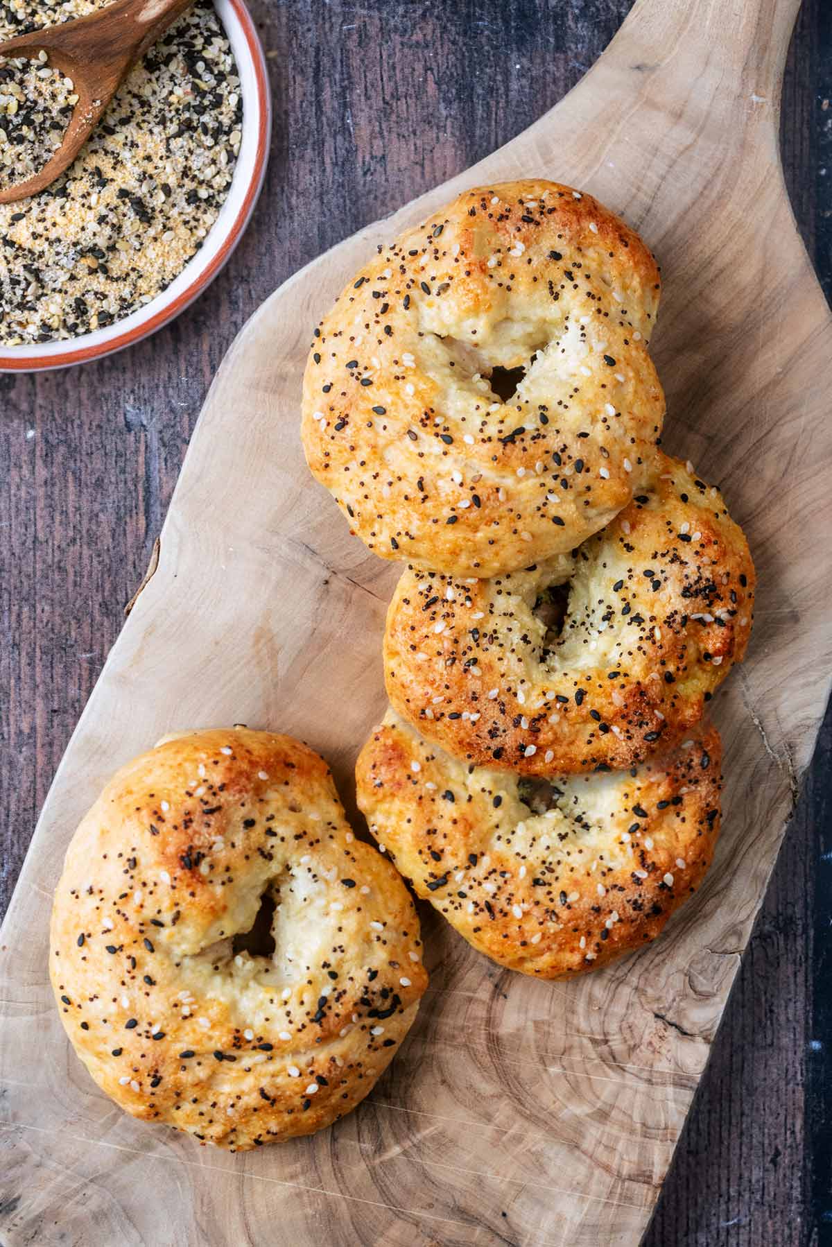 Four seeded bagels on a wooden board next to a bowl of seasoning.