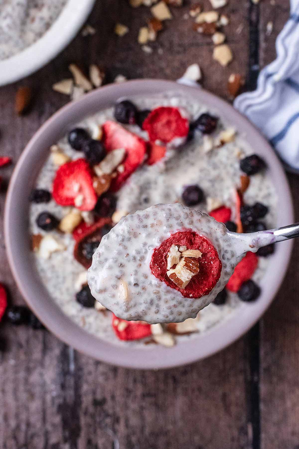 A spoon lifting some chia pudding, strawberry slice and crushed almond from a bowl.