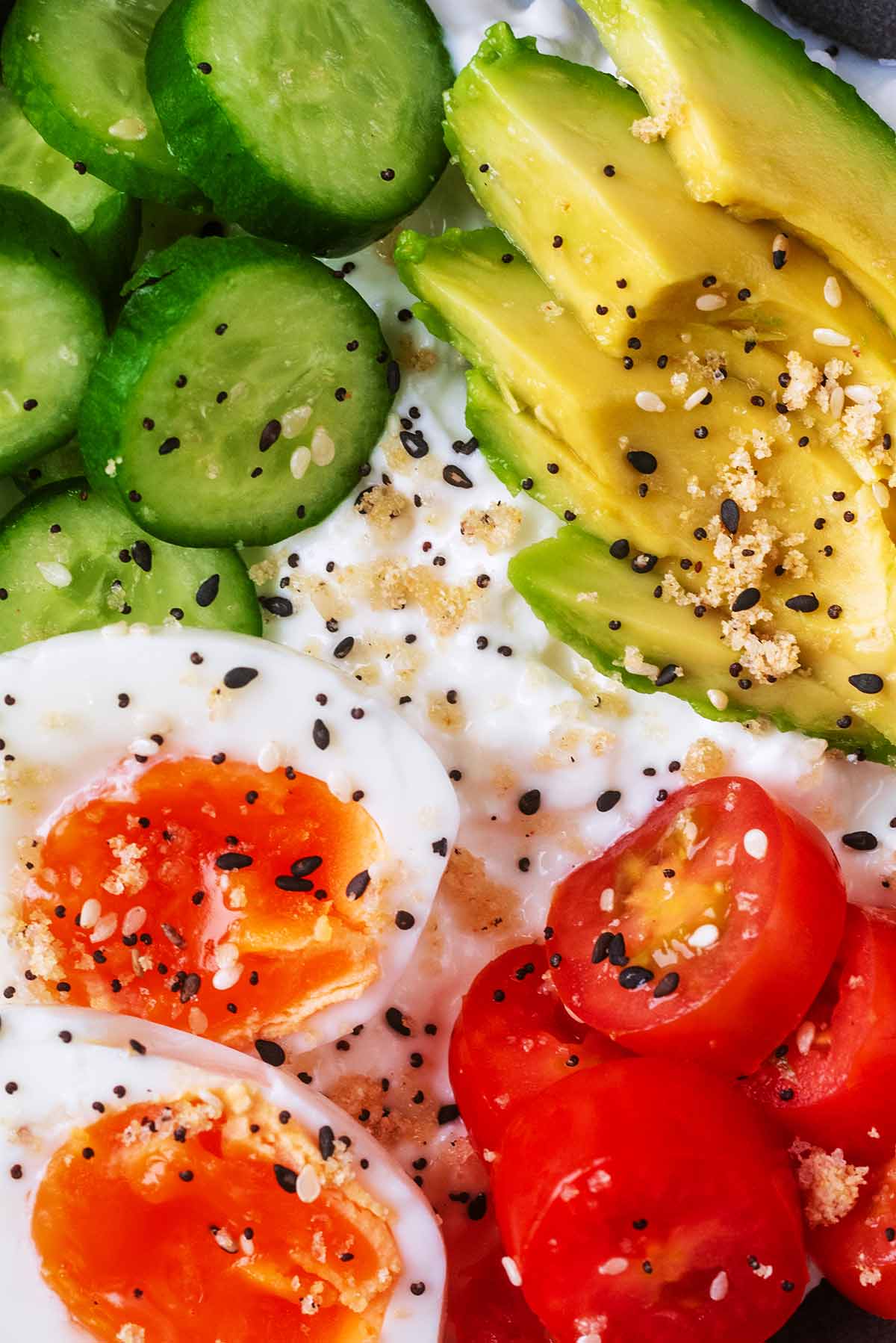 A boiled egg cut in half next to avocado, tomatoes and cucumber on top of cottage cheese.