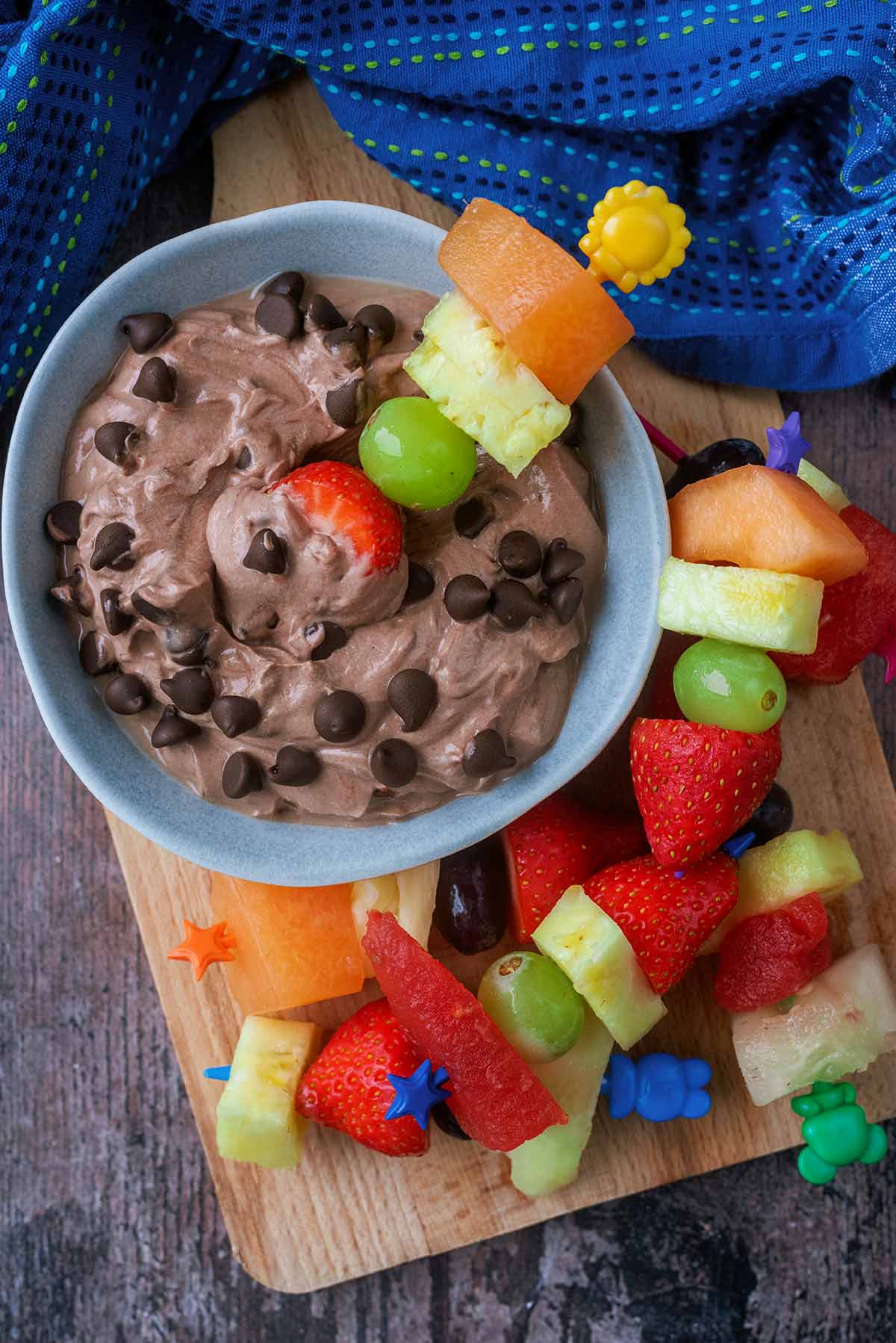 A bowl of chocolate dip with a fruit kebab in it and more fruit kebabs nearby.