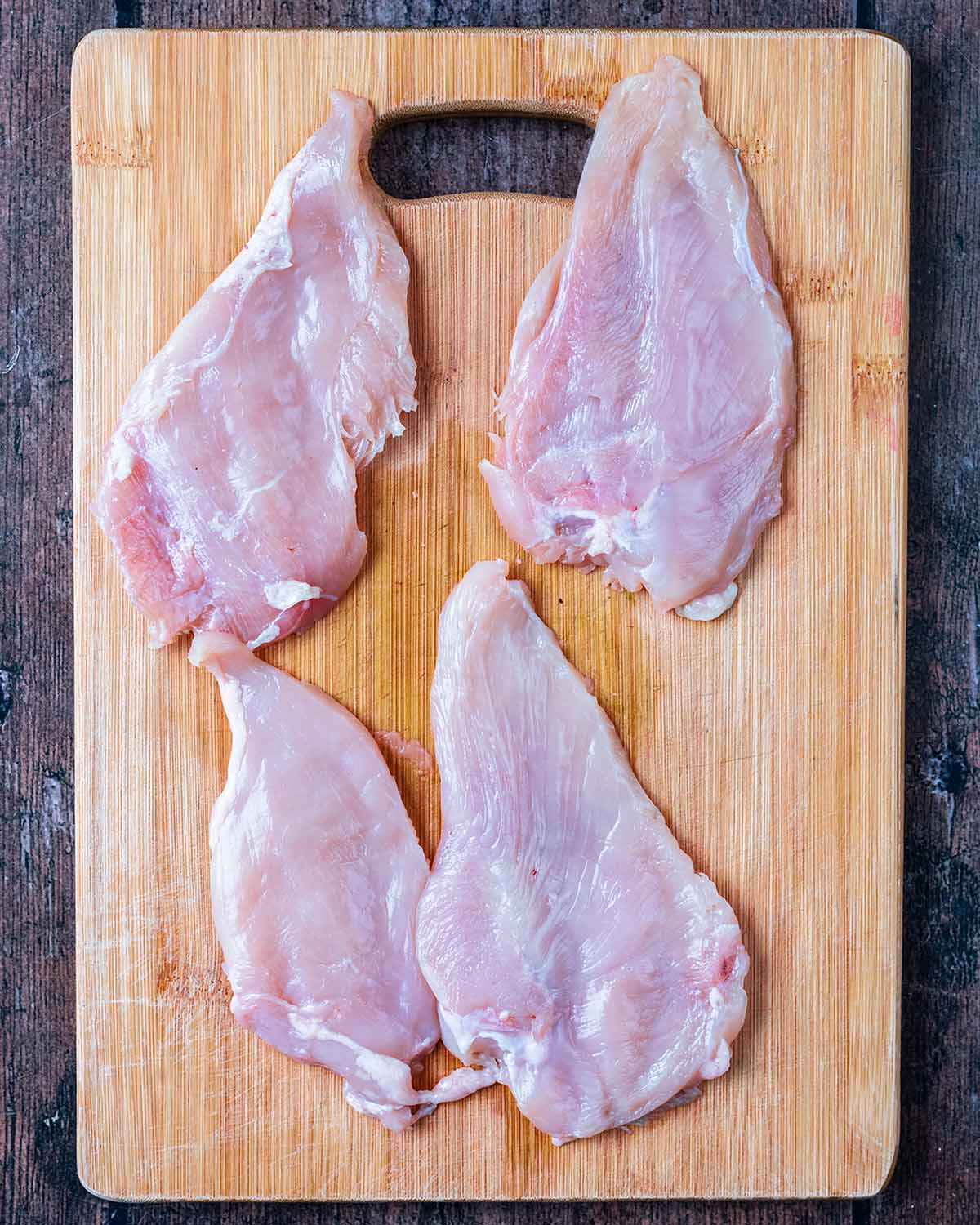 Four pounded chicken breasts on a wooden chopping board.