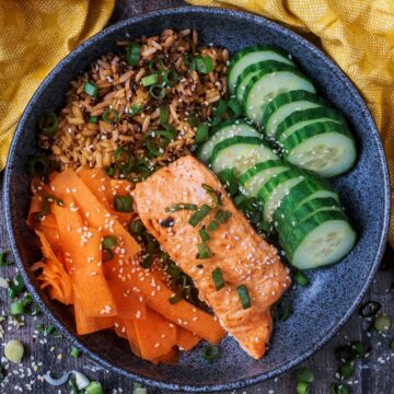 A bowl of bang bang salmon with rice, carrot and sliced cucumber.