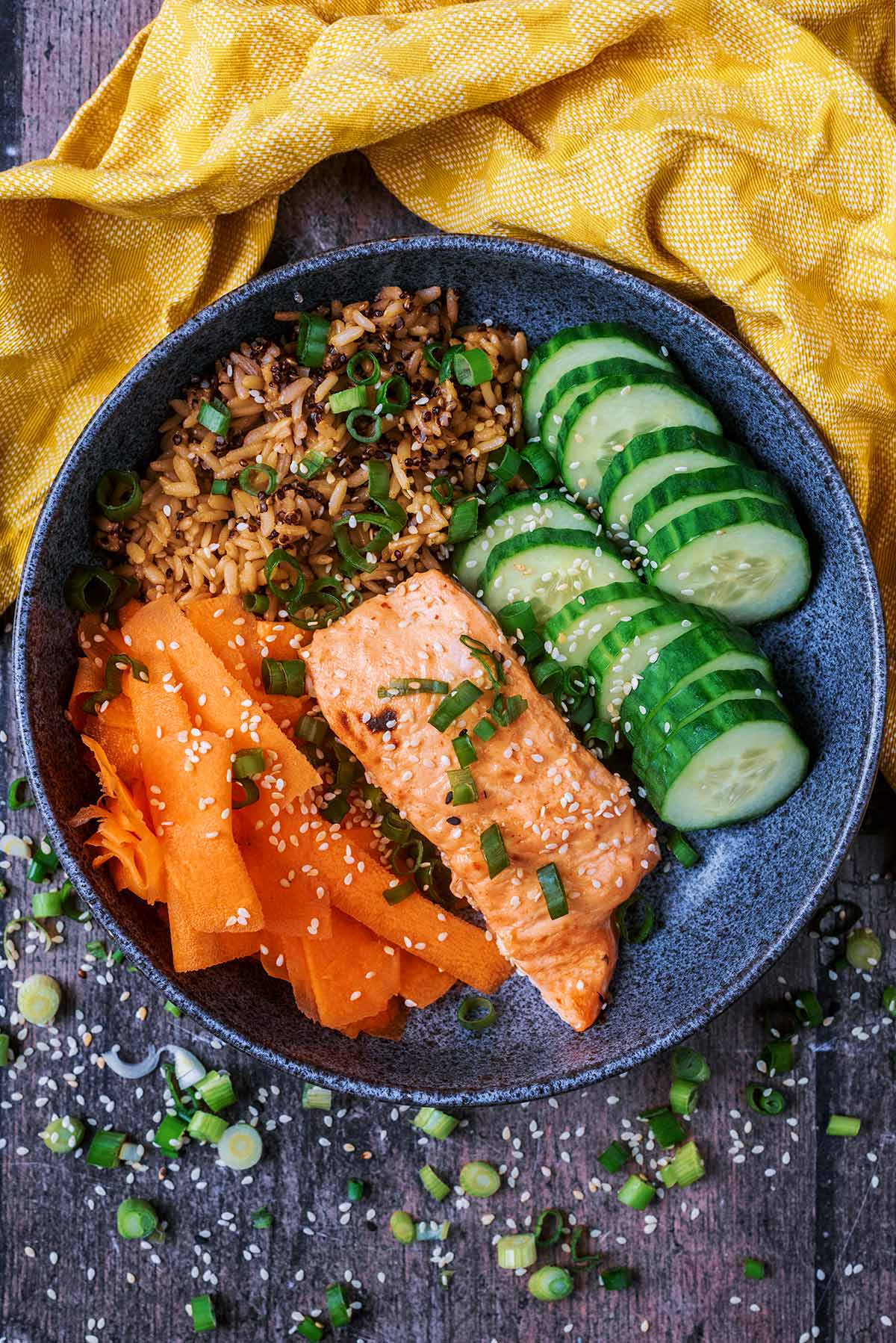 A bowl containing a cooked salmon fillet, some rice, ribboned carrot and sliced cucumber.