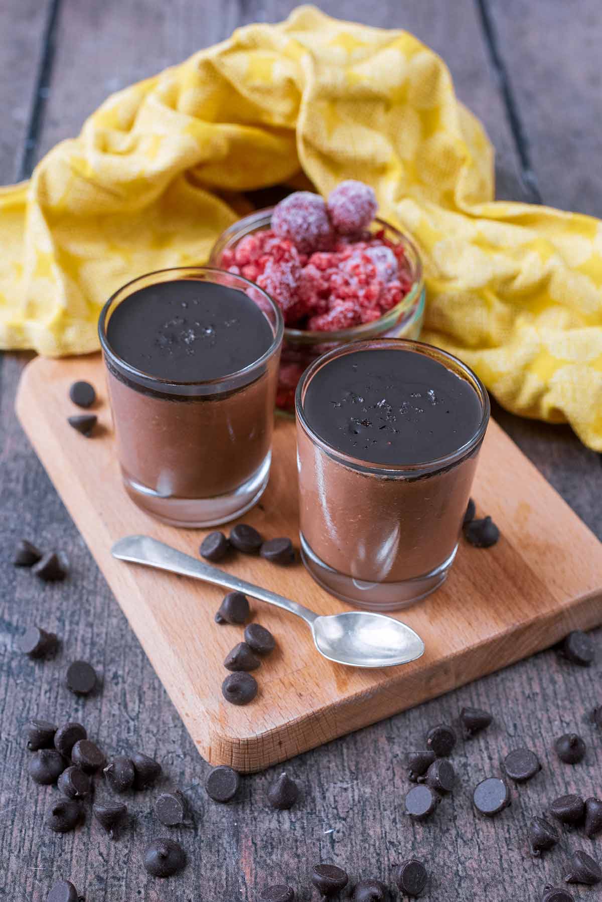 Glasses of chocolate mousse on a board next to some raspberries and chocolate chips.