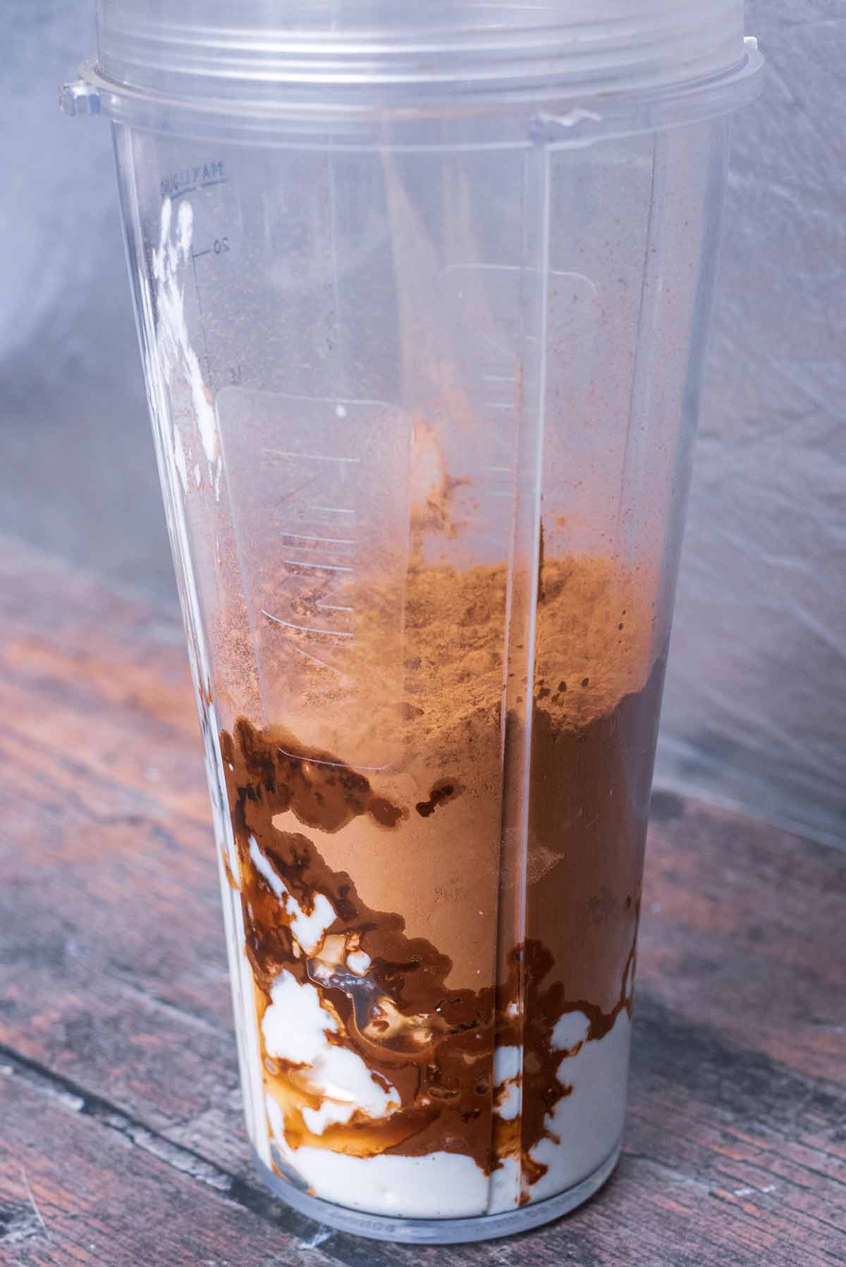 A blender jug containing cottage cheese, cacao powder, maple syrup and vanilla extract.