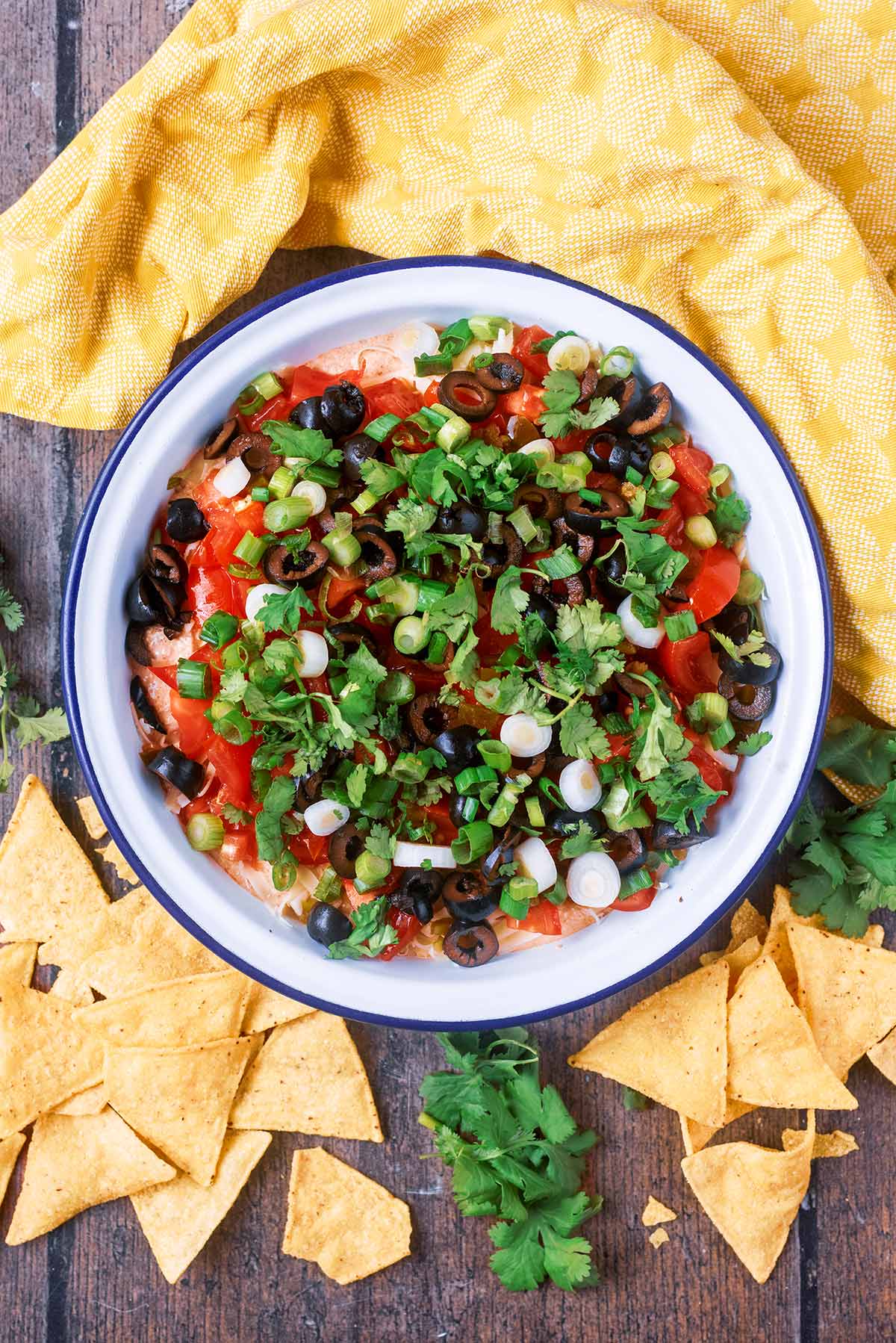 A bowl of taco dip surrounded by tortilla chips, cilantro leaves and a yellow towel.