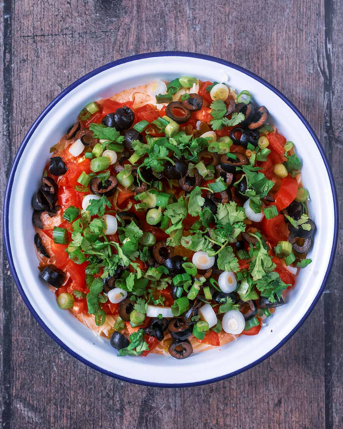 The bowl of dip topped with chopped tomatoes, jalapenos, olives and coriander leaves.