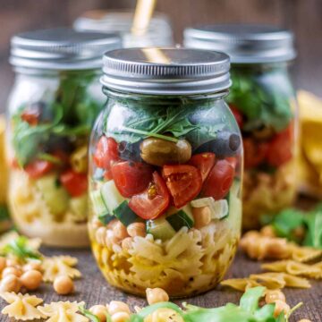 Pasta salad jars on a wooden surface surrounded by dry pasta and chickpeas.