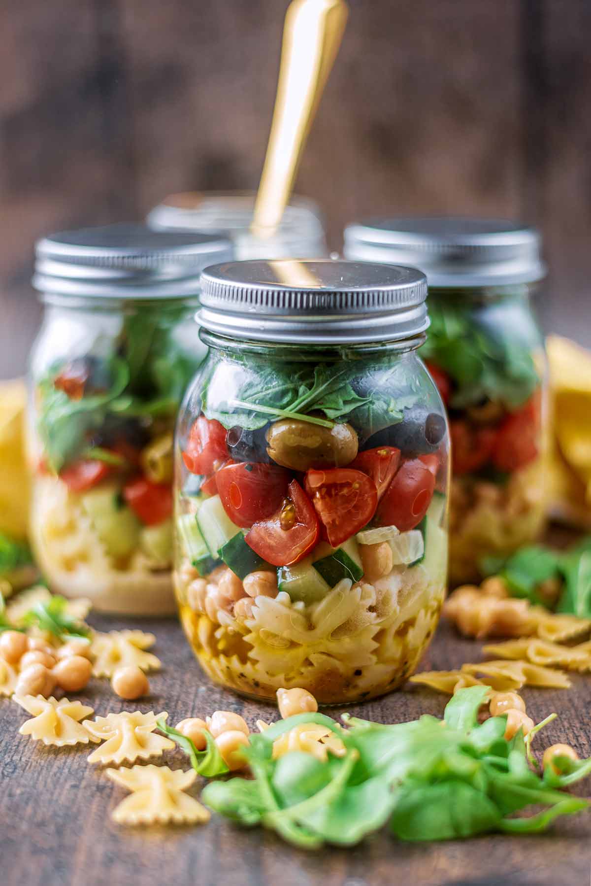 Four jars full of pasta salad. Lettuce, pasta and chickpeas surround the jars.