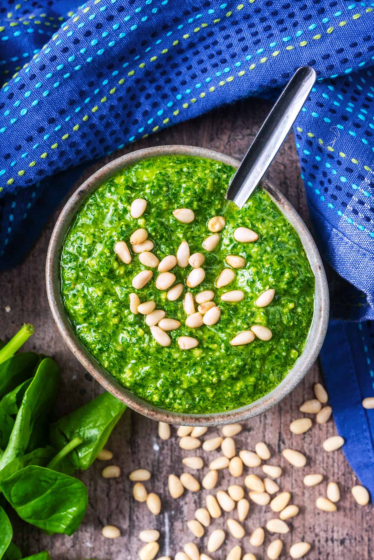 A bowl of pesto with a spoon next to a bunch of basil and some pine nuts.