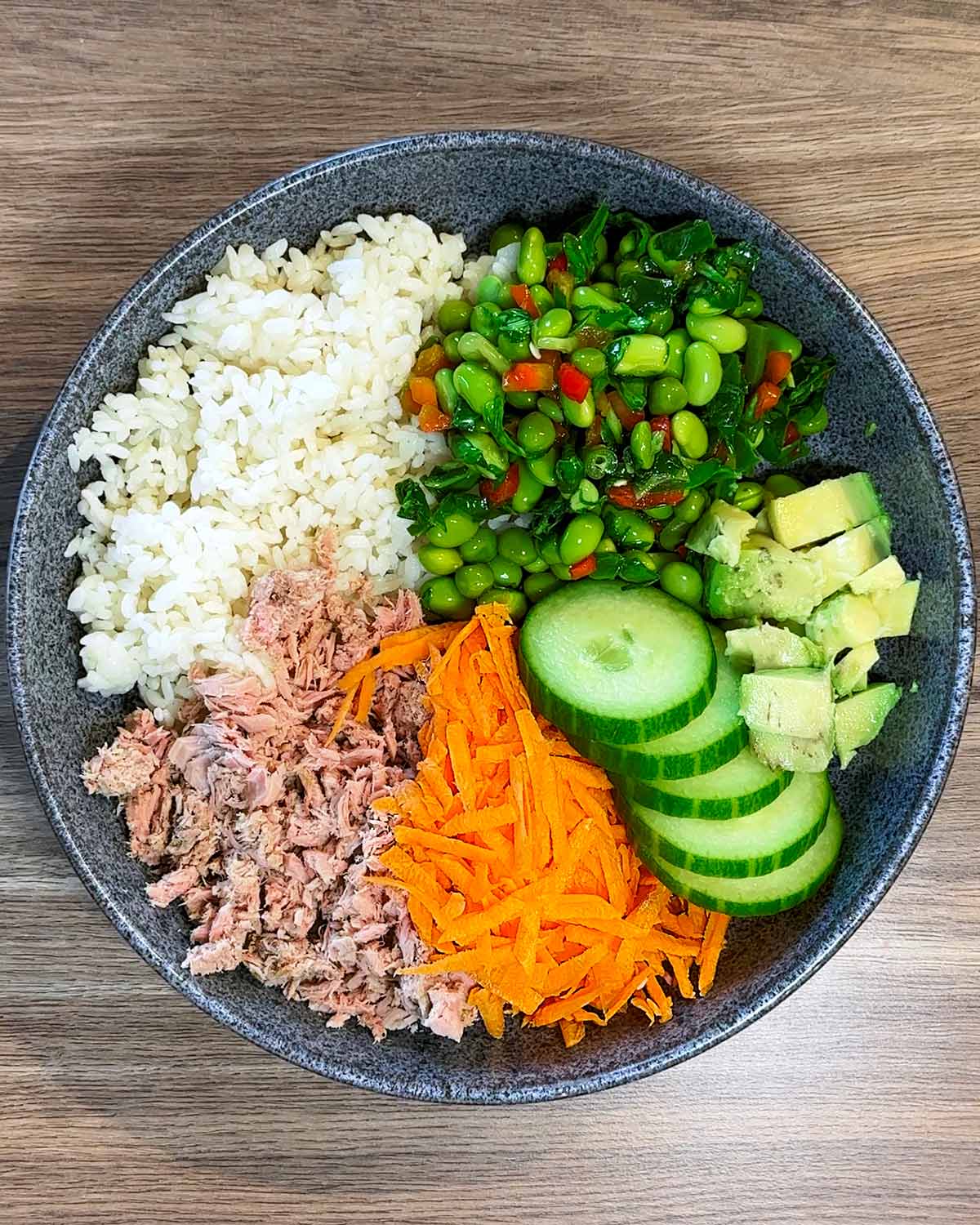 A bowl containing rice, tuna, sliced cucumber, grated carrot, avocado and edamame beans.