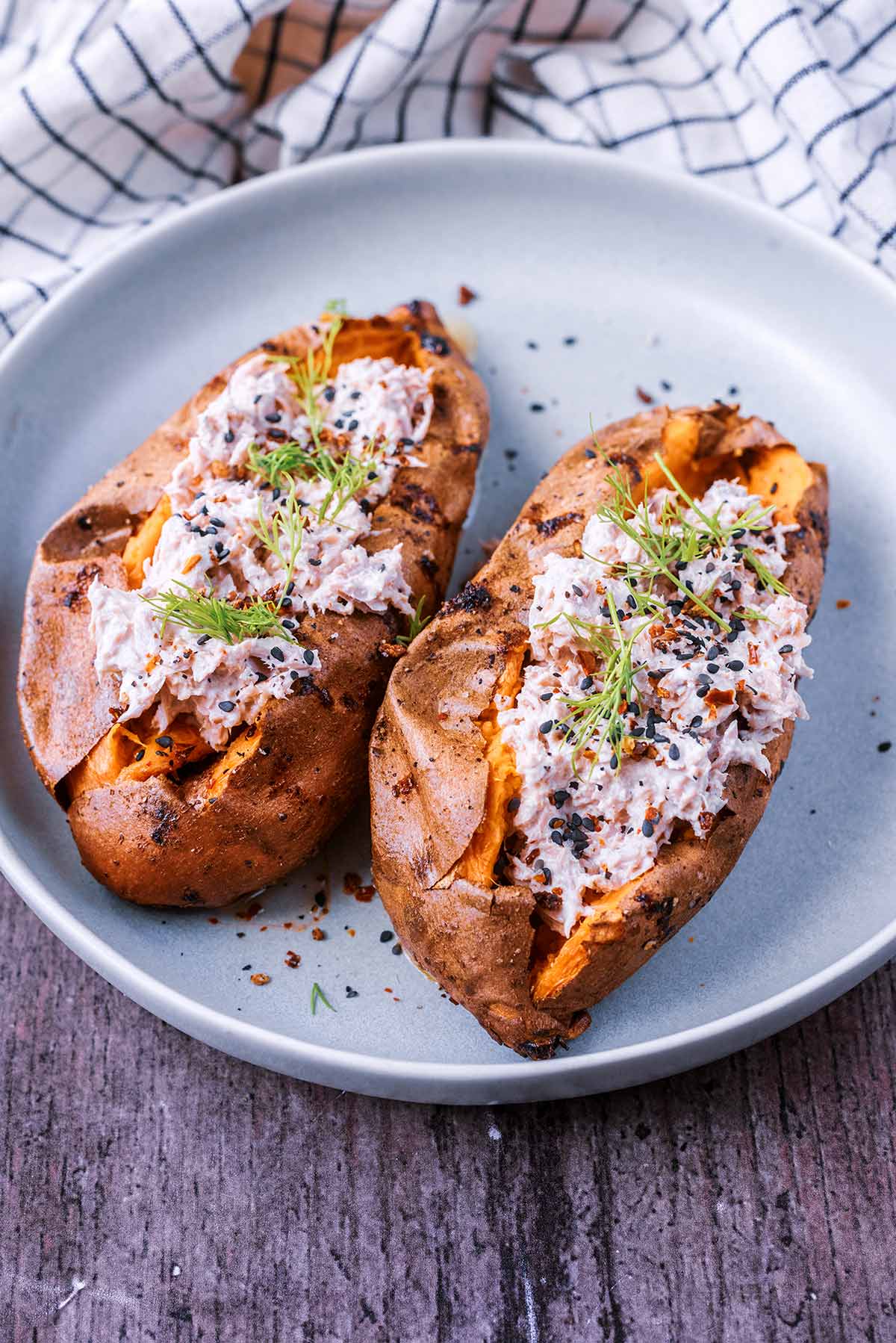 Two tuna mayonnaise topped sweet potatoes on a plate in front of a checked towel.