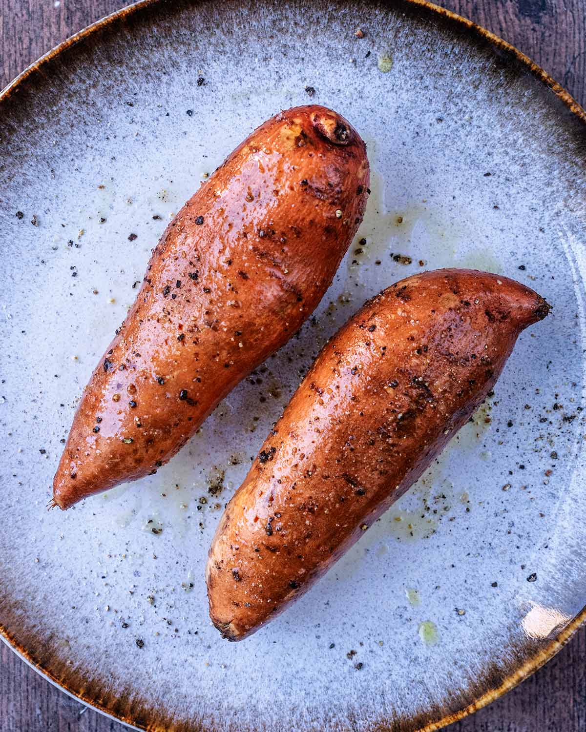 Two sweet potatoes on a plate coated in oil, salt and pepper.