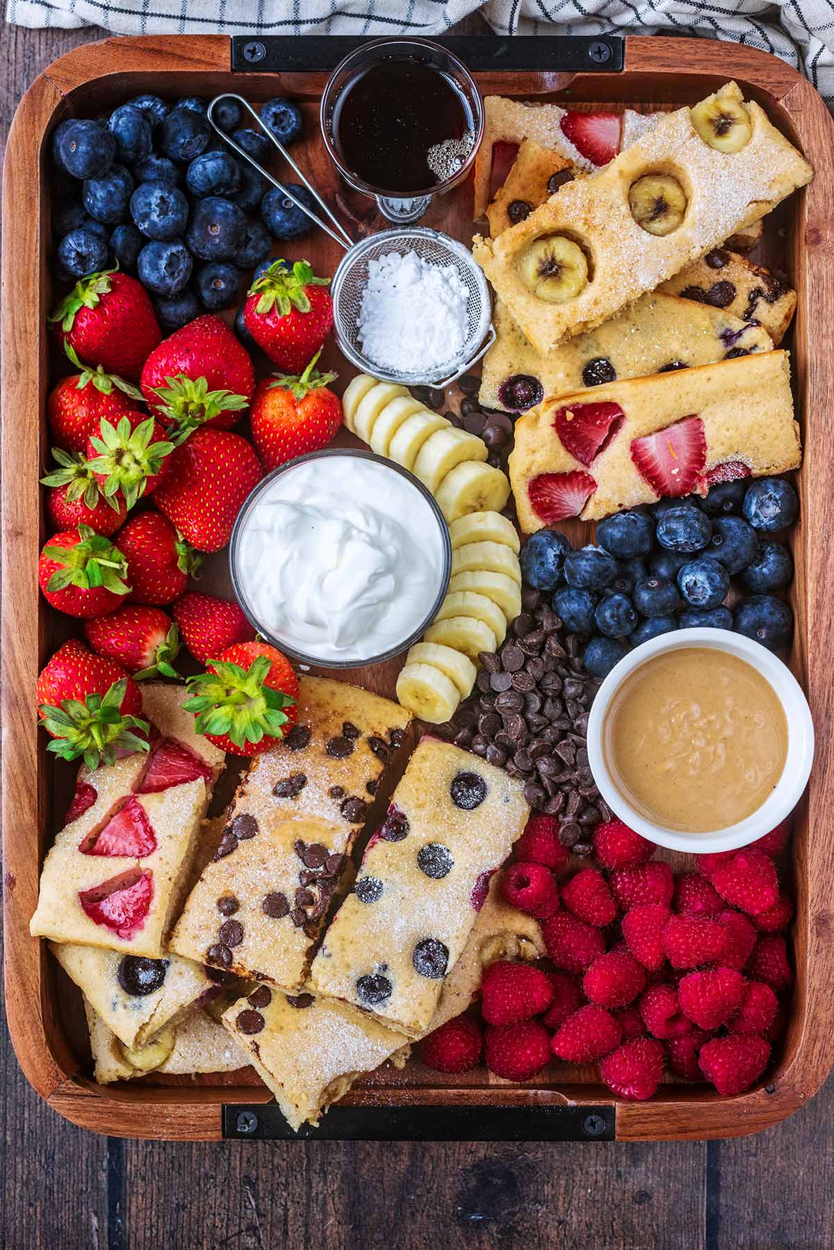 Slices of baked pancakes on a board with berries, chocolate chips, yogurt and peanut butter.