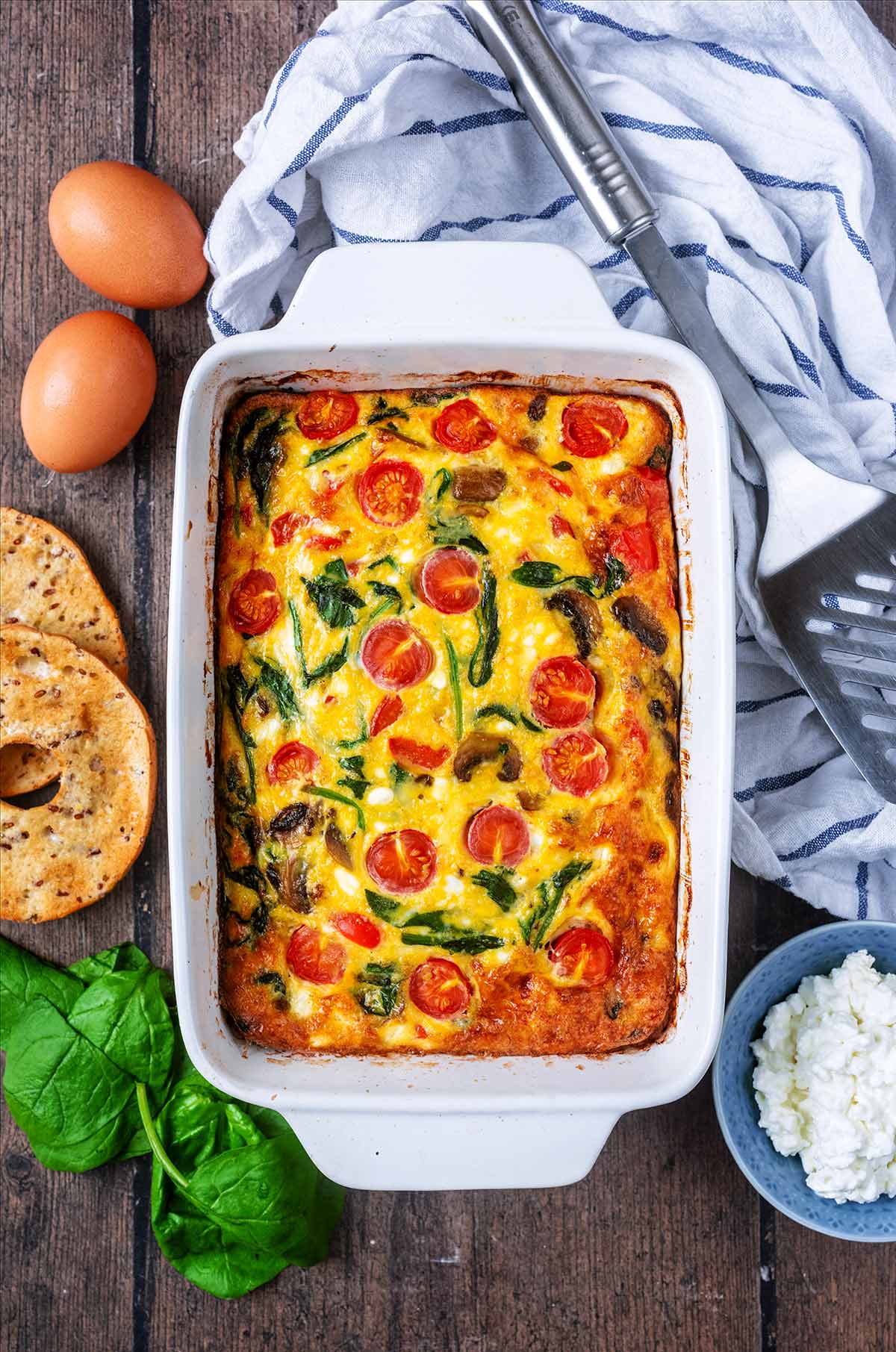 Frittata in a baking dish surrounded by spinach leaves, bagels a towel and some cottage cheese.