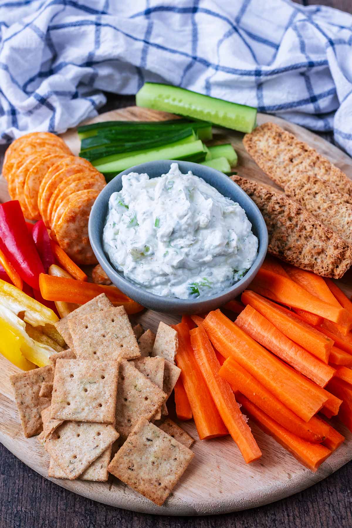 A bowl of creamy dip on a wooden board with carrot sticks, cucumber, bread and crackers.