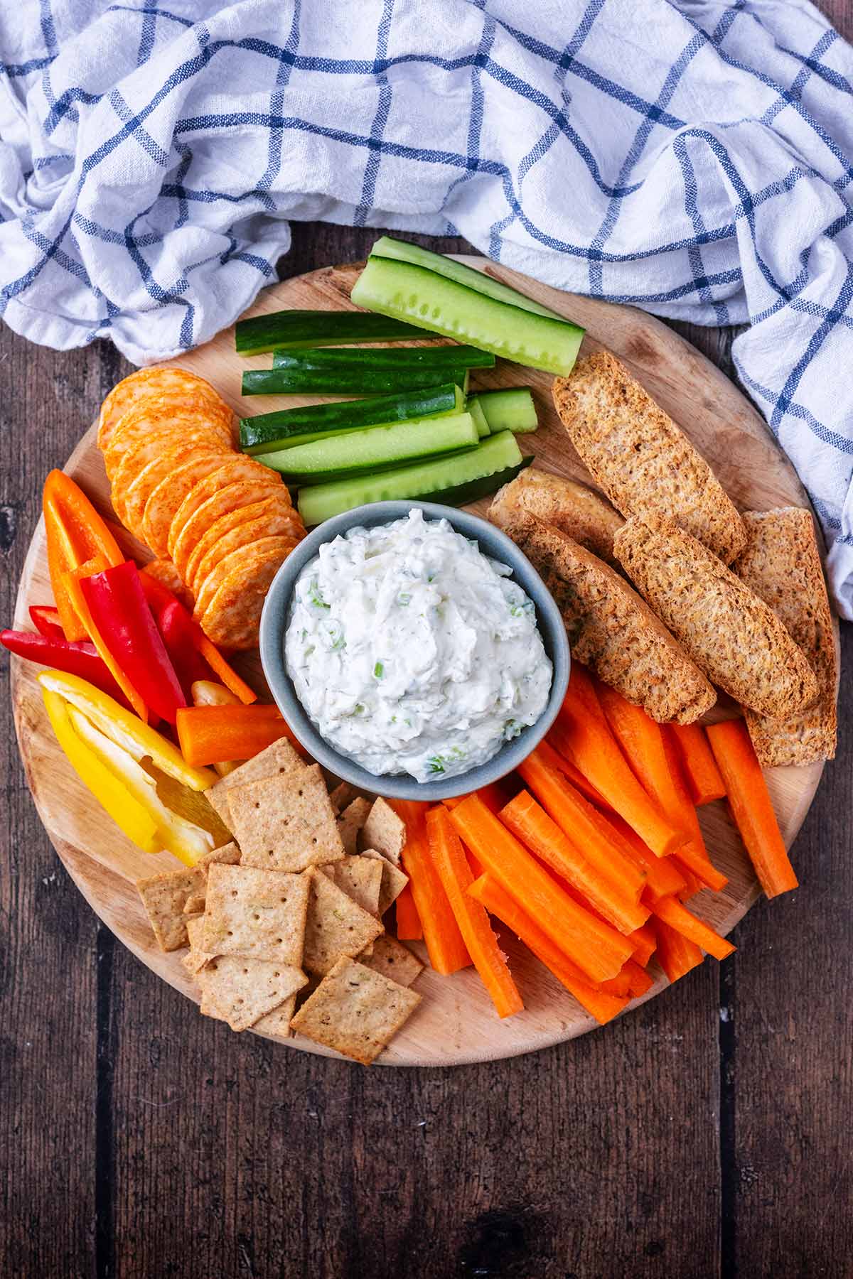 A wooden serving board with ranch dip, vegetable sticks, bread and crackers.