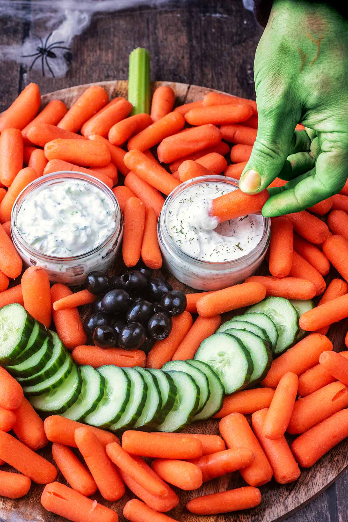 A green coloured hand dipping a carrot into some tzatziki.