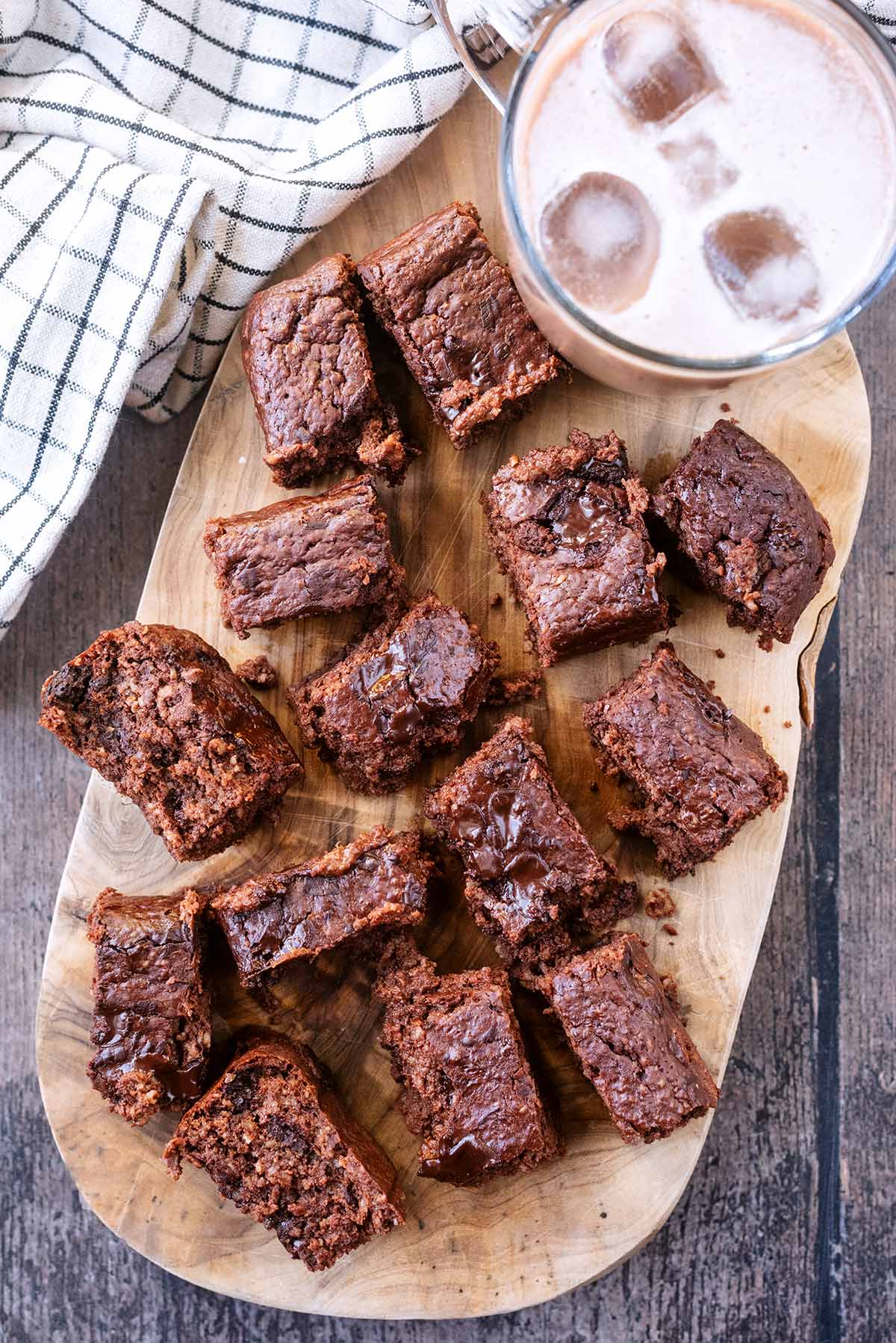 Fourteen small chocolate brownies on a wooden serving board with a cup of iced coffee.