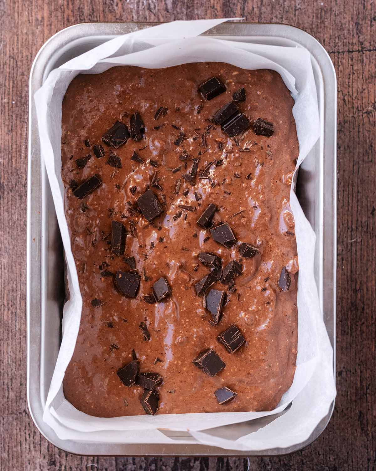 Brownie batter in a lined baking dish topped with small chunks of chocolate.