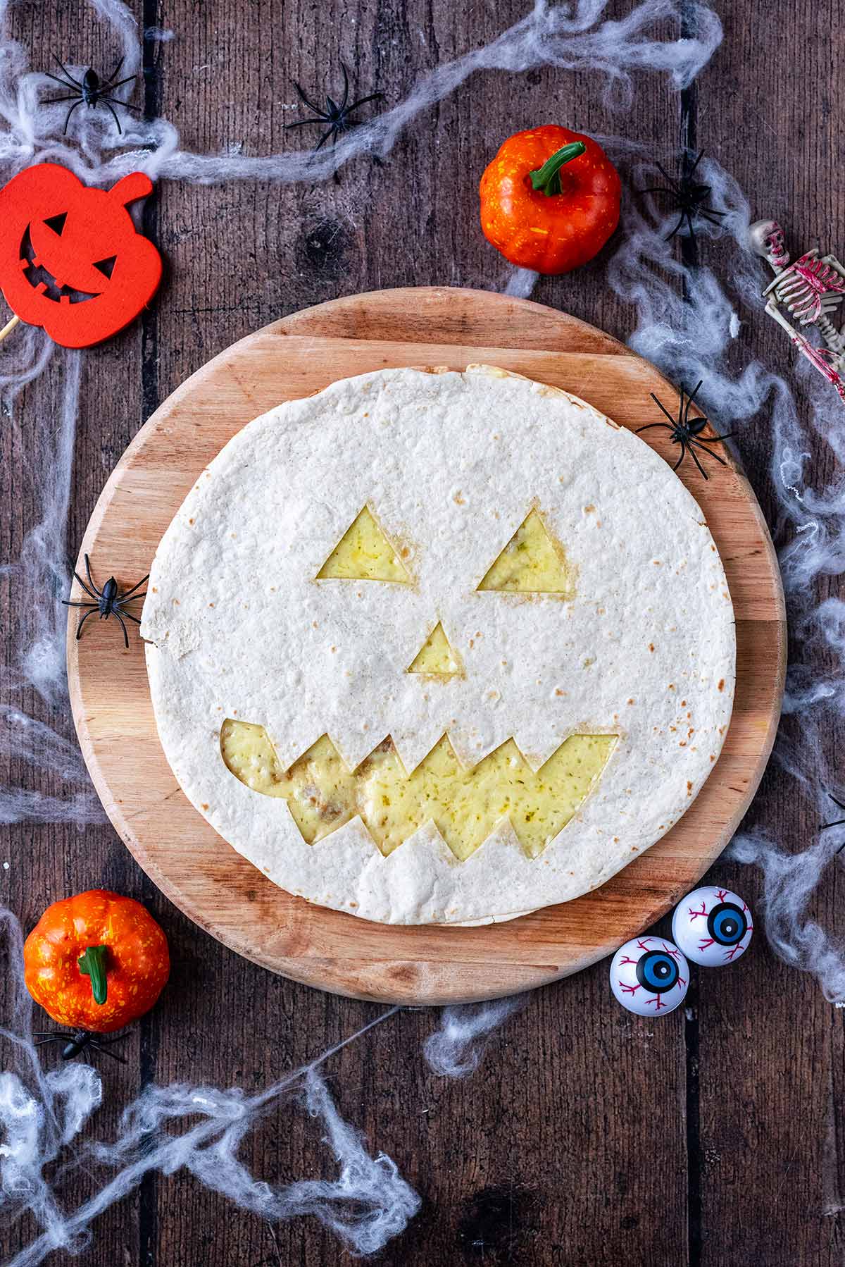 A quesadilla with cut out shapes like a Jack O'Lantern, surrounded by fake pumpkins and cobwebs.