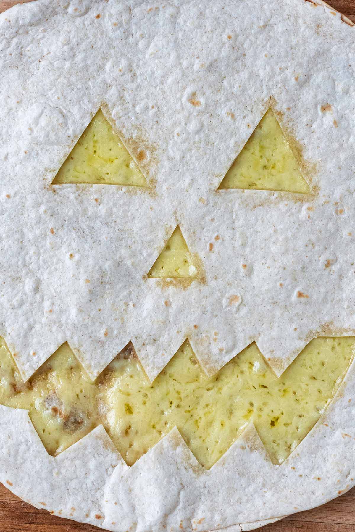 Melted cheese behind a flour tortilla that has been cut into a Jack O'Lantern design.