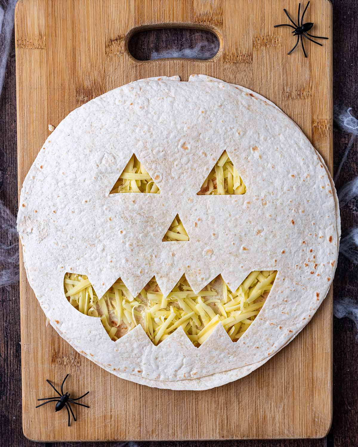 A Jack O'Lantern tortilla on top of a cheese loaded other tortilla.