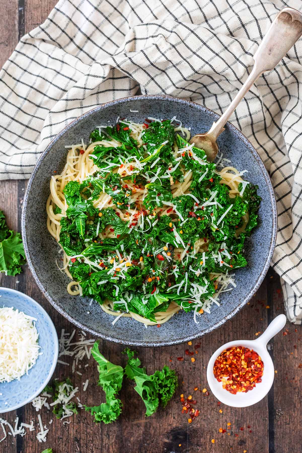 A bowl of cooked kale and spaghetti on a wooden surface with Parmesan and chilli flakes.