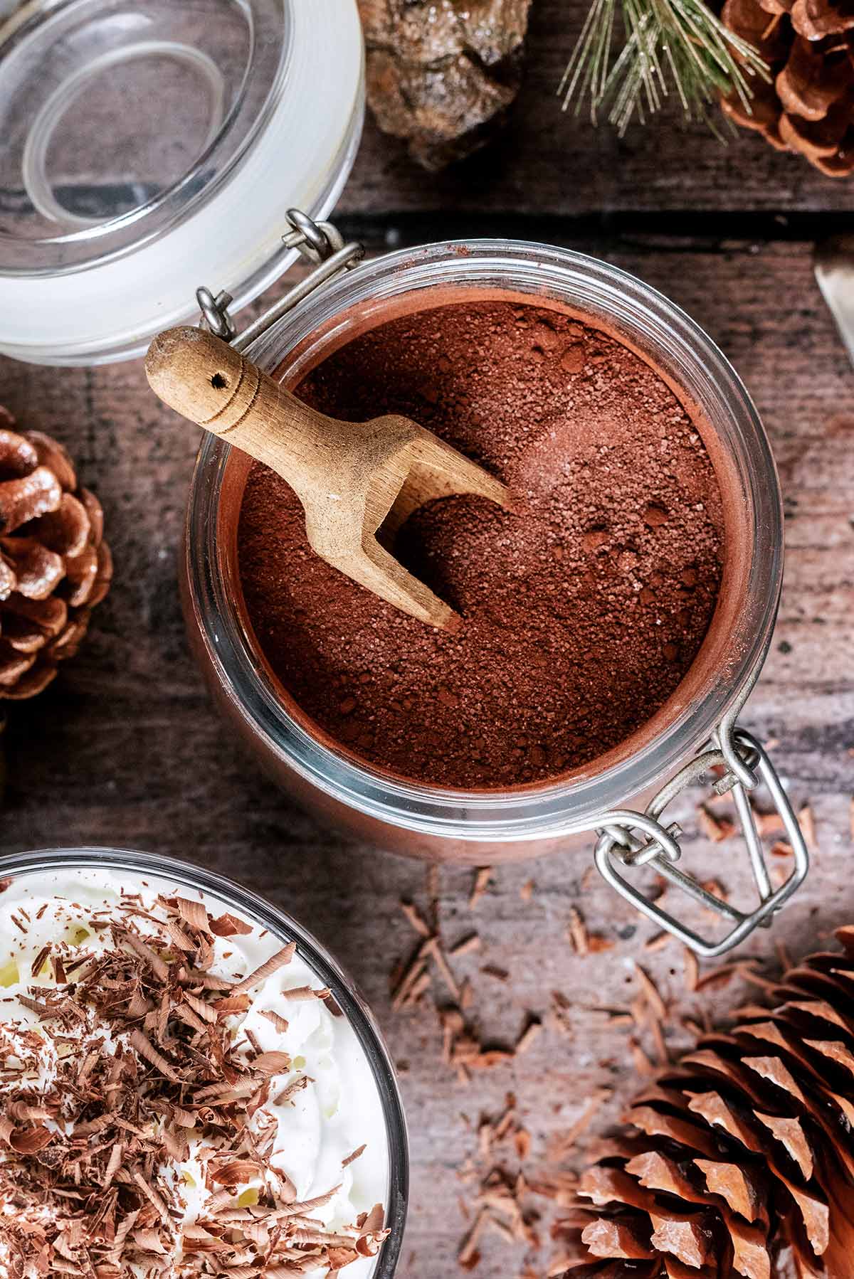 A jar of hot chocolate powder next to a mug of hot chocolate and pine cones.