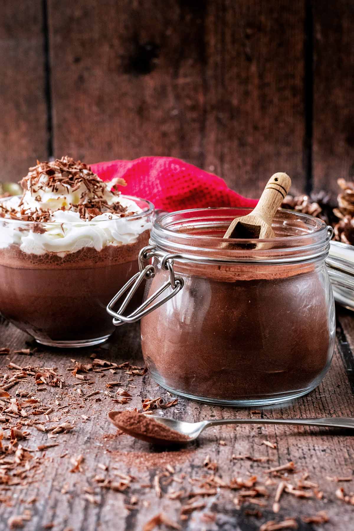 A jar of hot chocolate mix in front of a whipped cream topped hot chocolate drink.