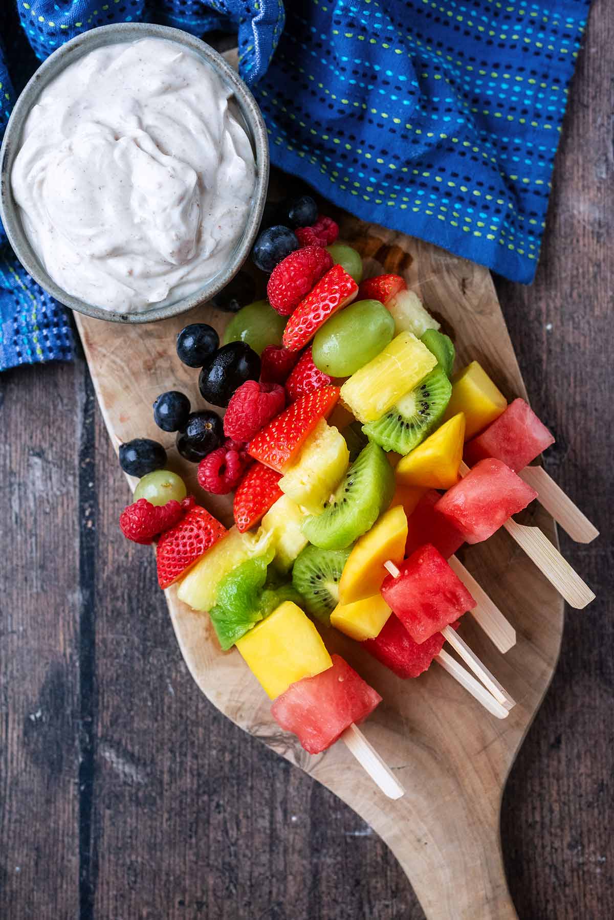 Skewers with chunks of fruit on them on a board with a creamy dip.