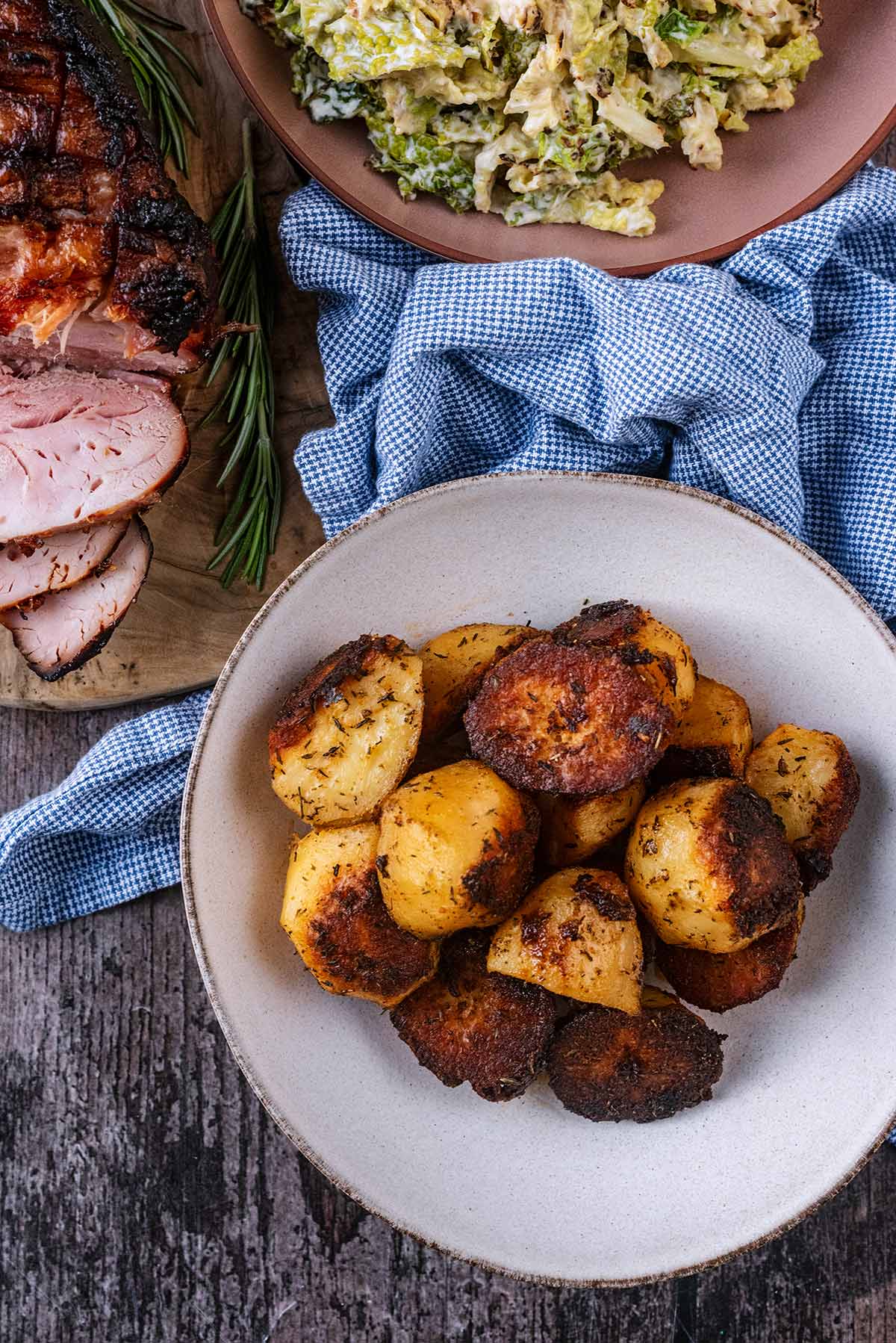 A bowl of roast potatoes next to a joint of ham and a bowl of cooked cabbage.