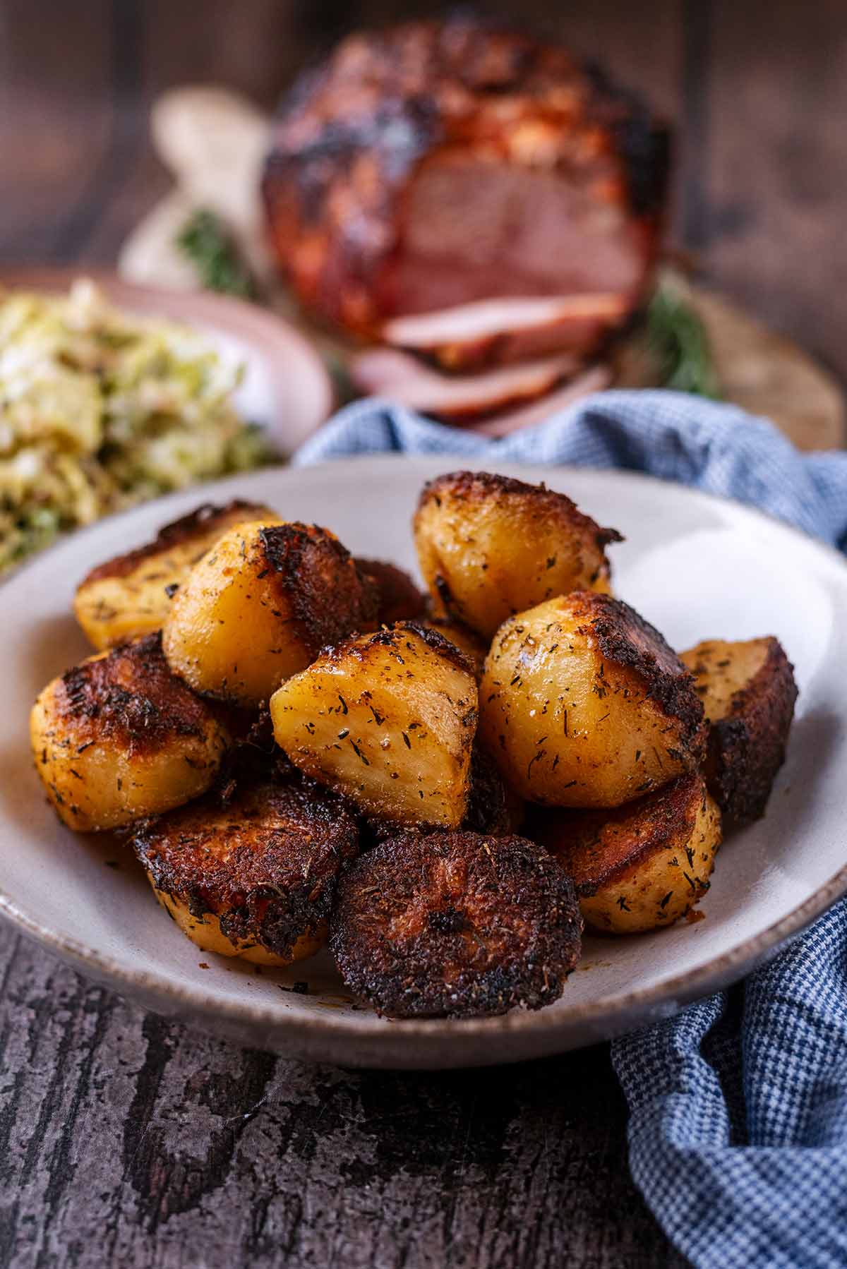 Roast potatoes on a plate with sliced ham and a bowl of vegetables in the background.