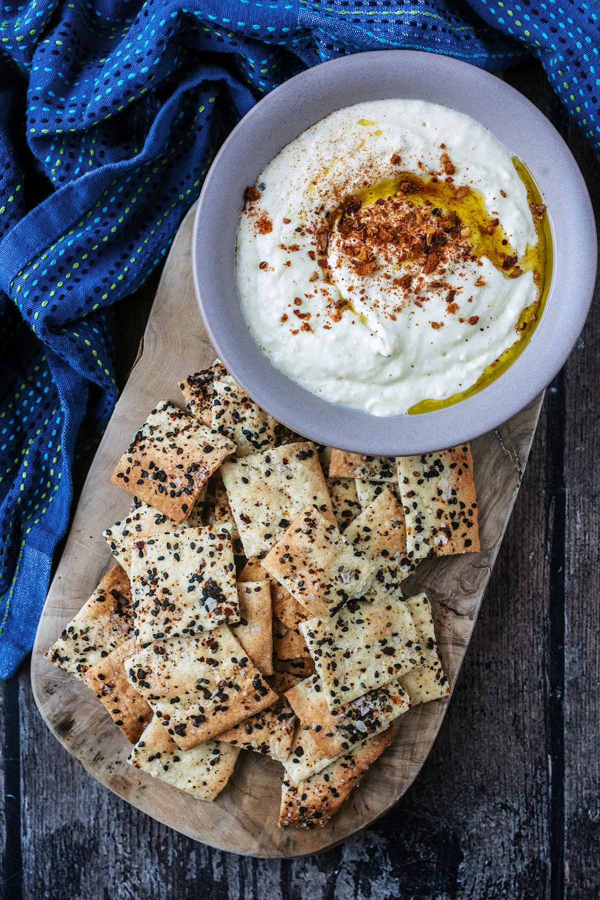 a wooden serving board with a pile of small square crackers and a bowl of hummus.