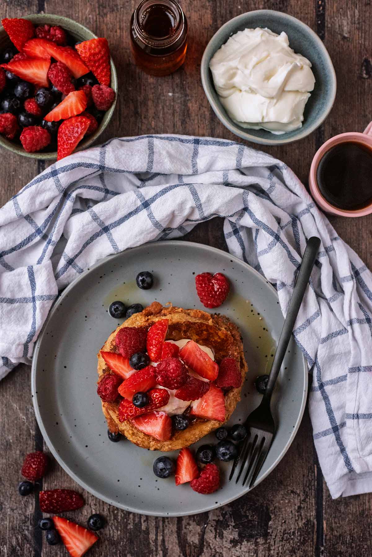 A plate of pancakes next to a towel, bowl of berries, some yogurt and a cup of coffee.