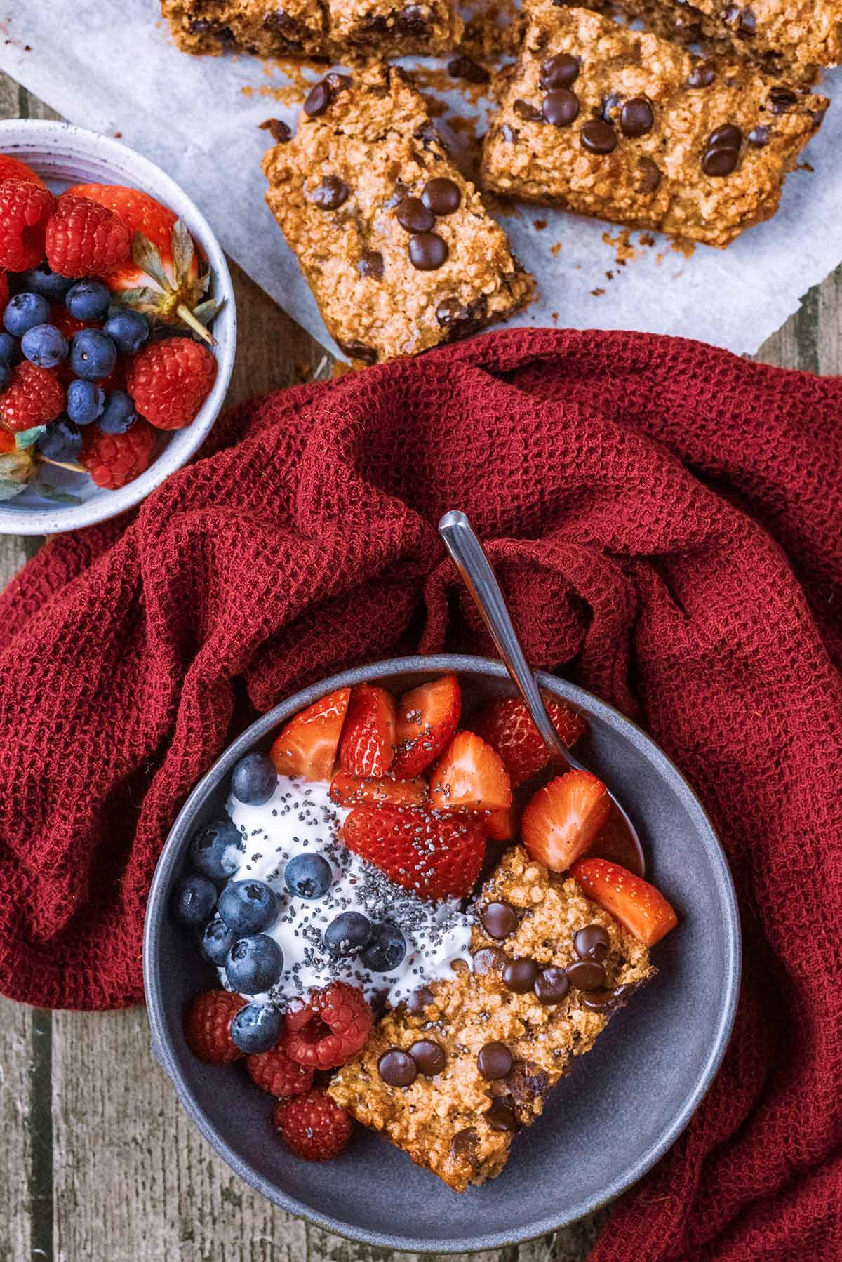 A bowl containing oatmeal cake, yogurt and berries next to a towel and some more berries.