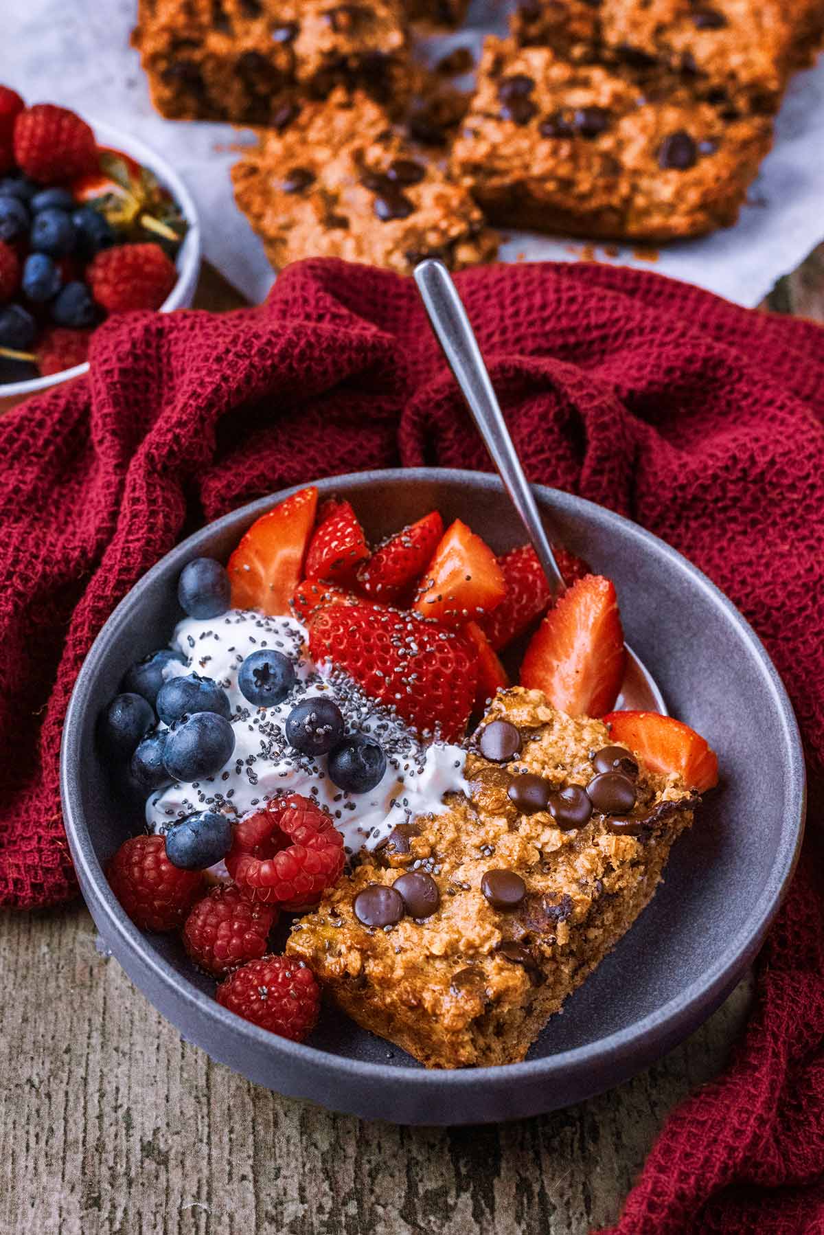 A bowl of oatmeal cake, yogurt and berries in front of a red towel and more of the cake.