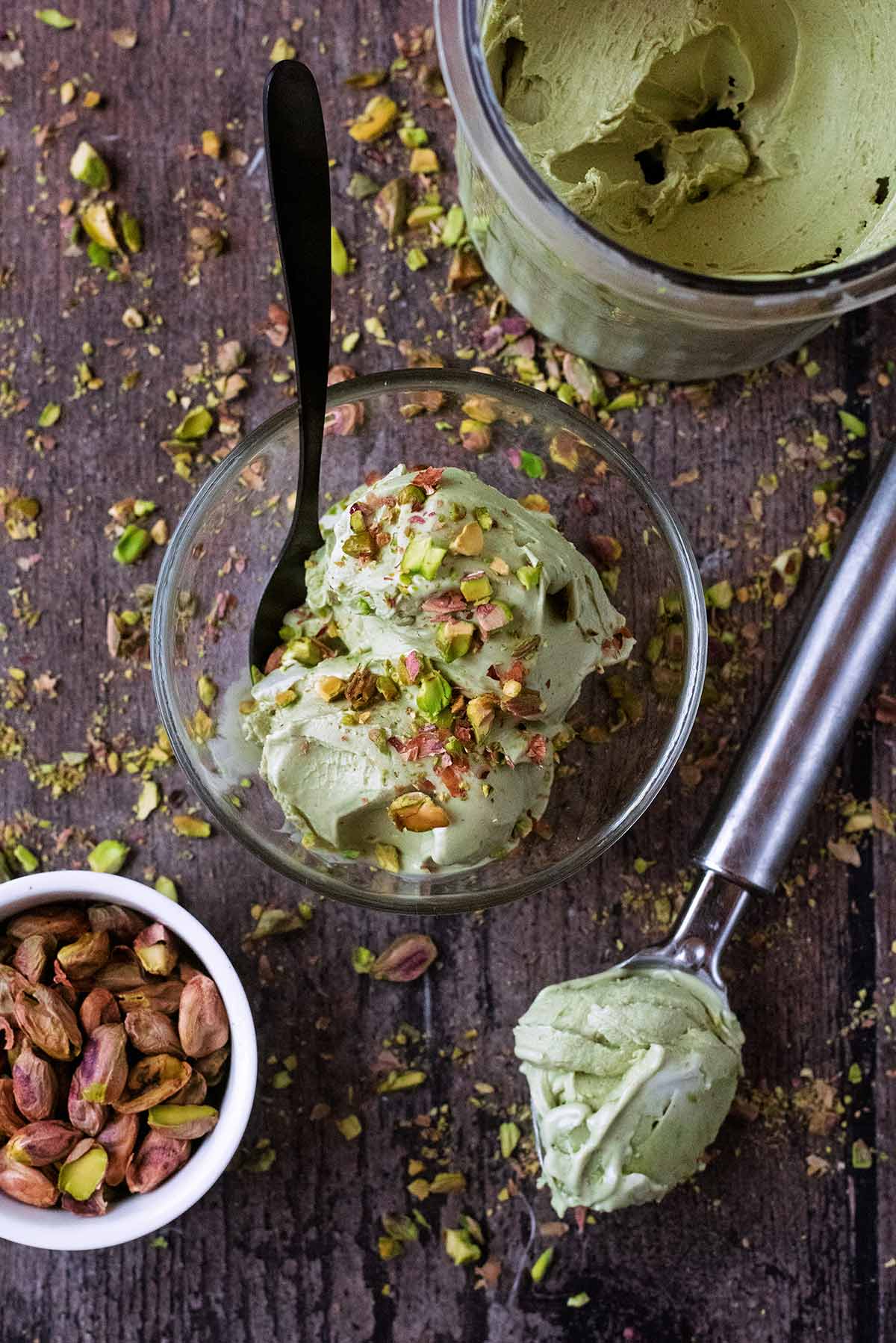 a bowl of pistachio ice cream next to a tub of ice cream, scoop and bowl of pistachios.