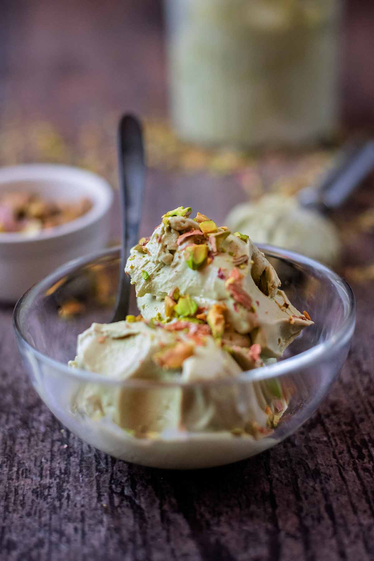 A bowl of pistachio ice cream topped with crushed pistachios, in front of a tub of more ice cream.