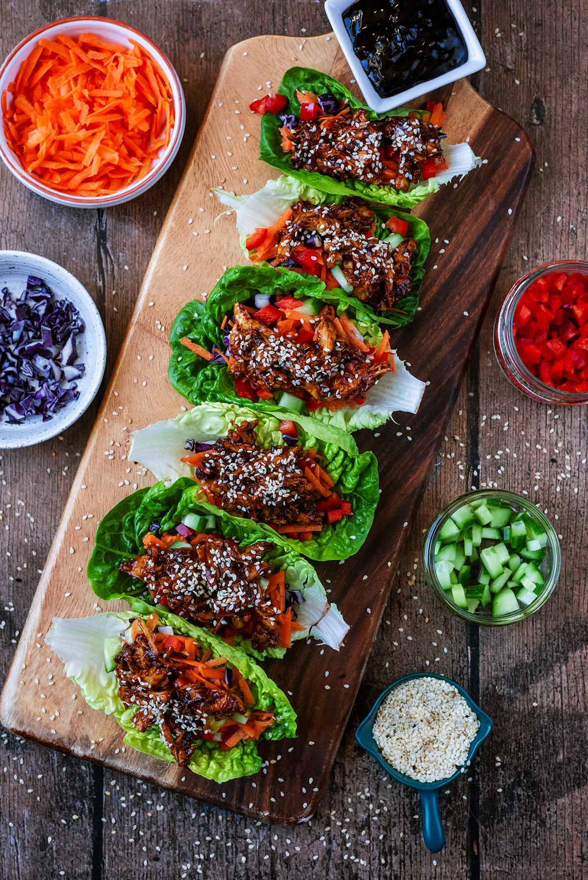 Six chicken lettuce wraps on a large serving board surrounded by bowls of chopped vegetables.