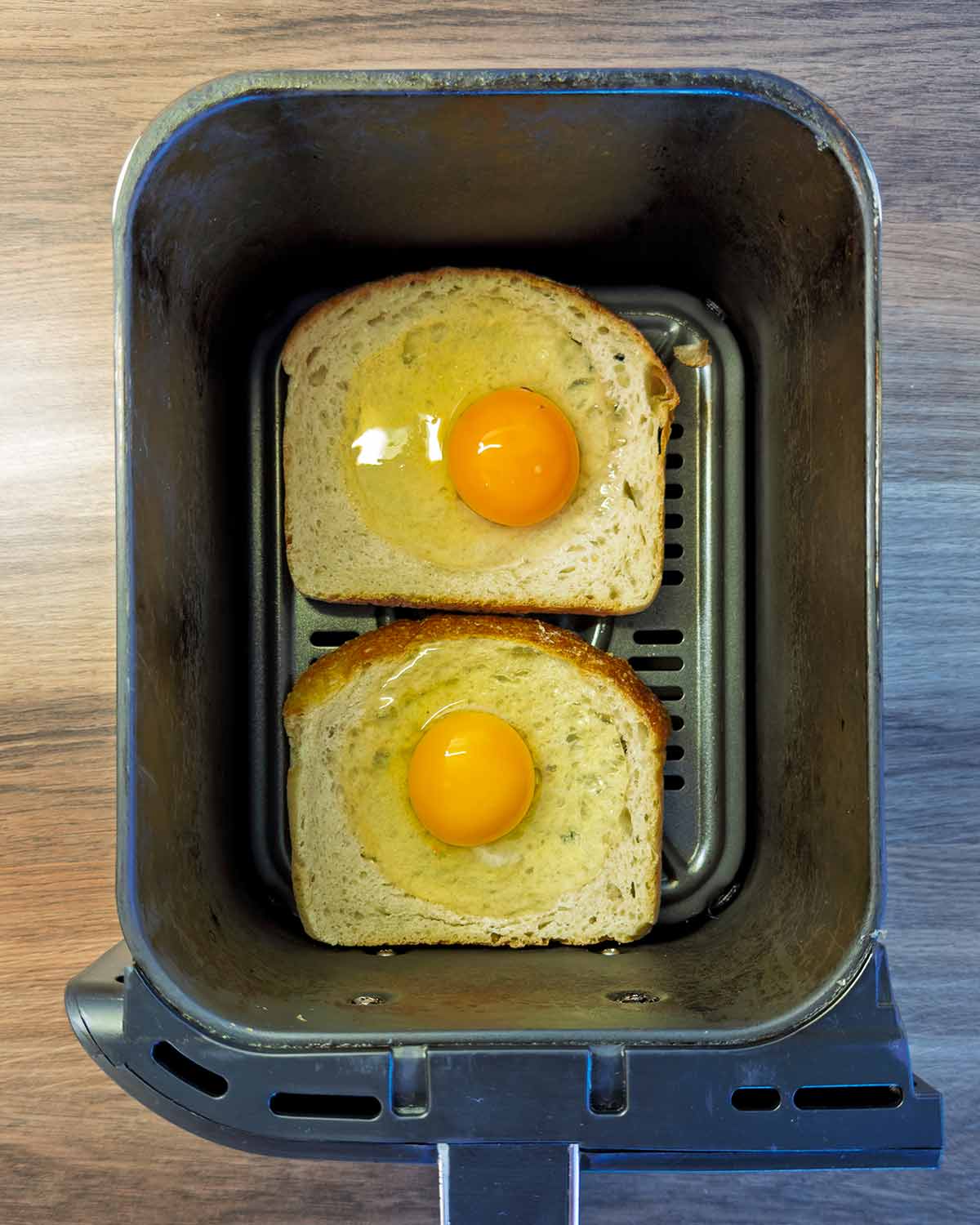 The bread in an air fryer basket with an egg cracked into each one.