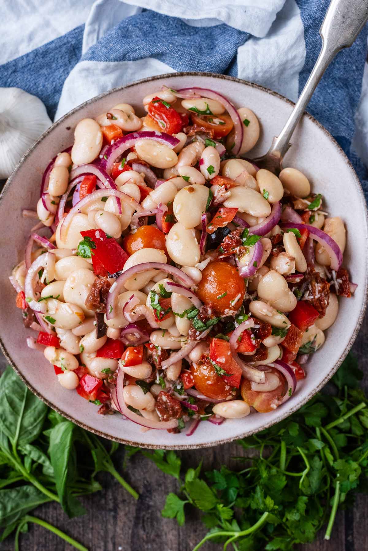 A bowl of bean salad next to bunches of herbs and a bulb of garlic.