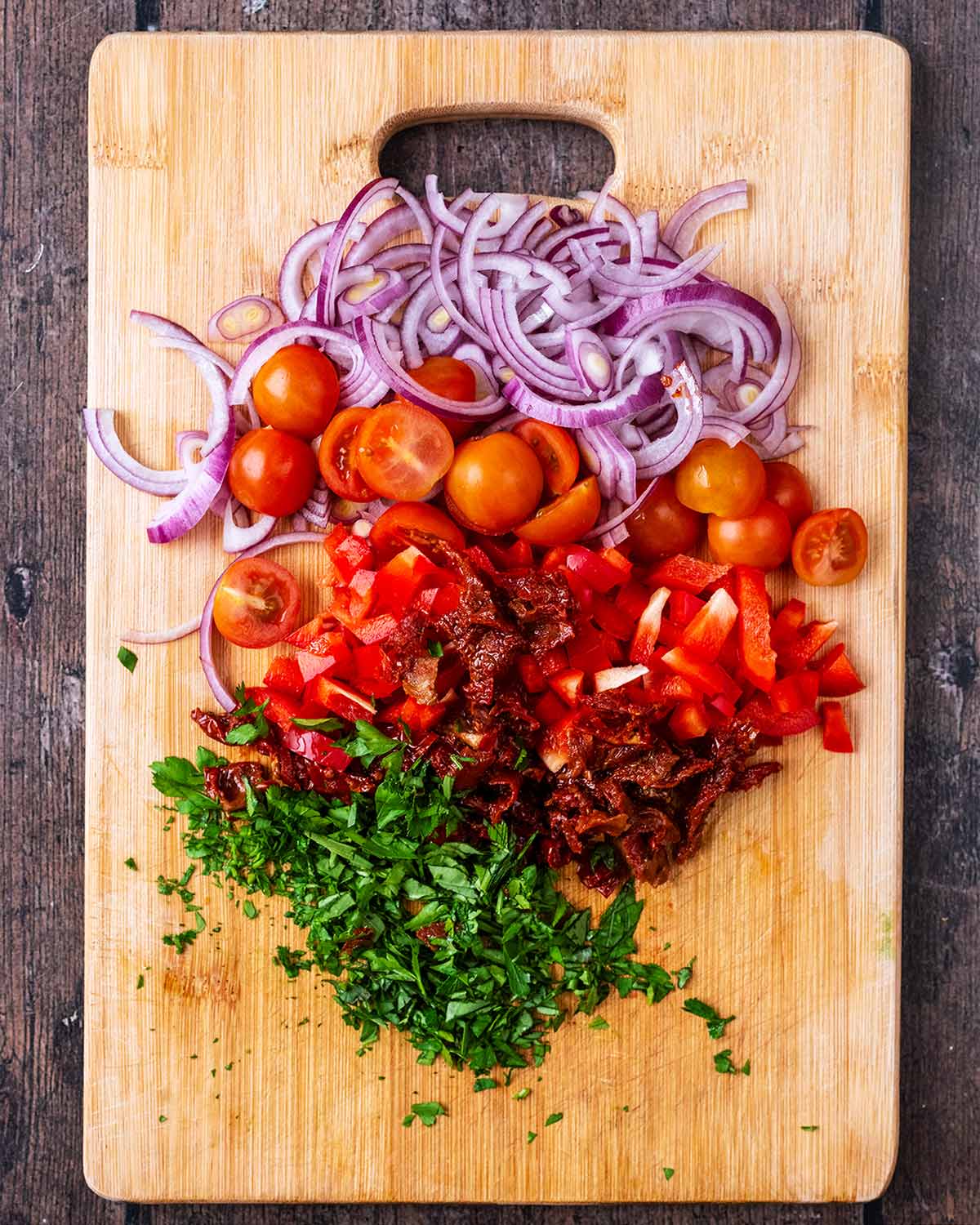 A wooden chopping board with chopped onions, tomatoes, bell pepper and herbs on it.