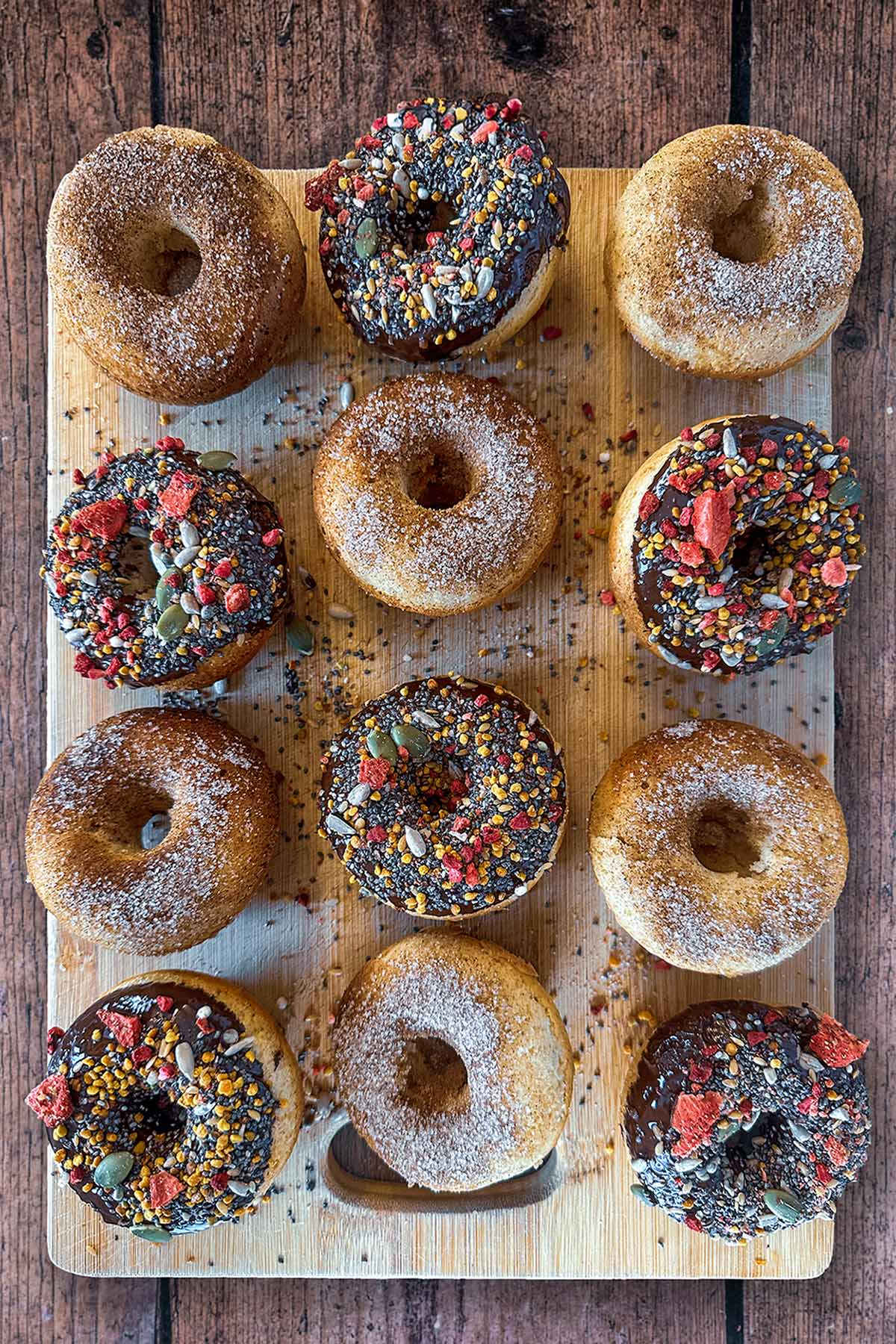 Twelve doughnuts on a wooden board, six of them topped with sugar, six topped with chocolate and sprinkles.