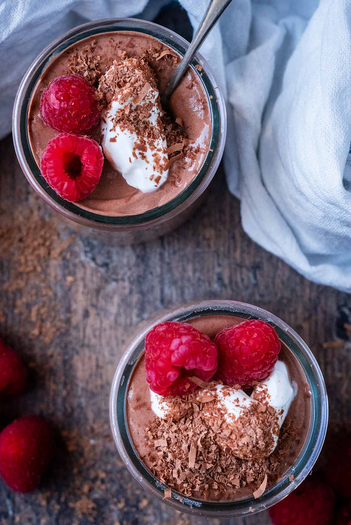 Two chocolate pudding pots as viewed from above. Both are topped with raspberries and yogurt.