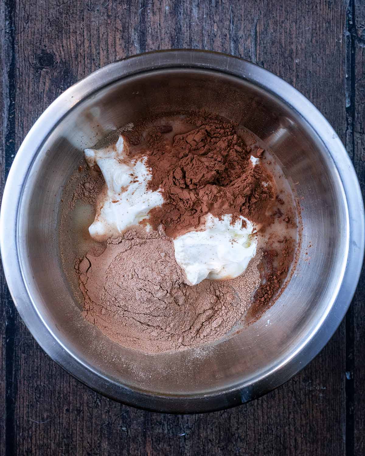 A mixing bowl containing yogurt, cacao powder, protein powder, milk and maple syrup.