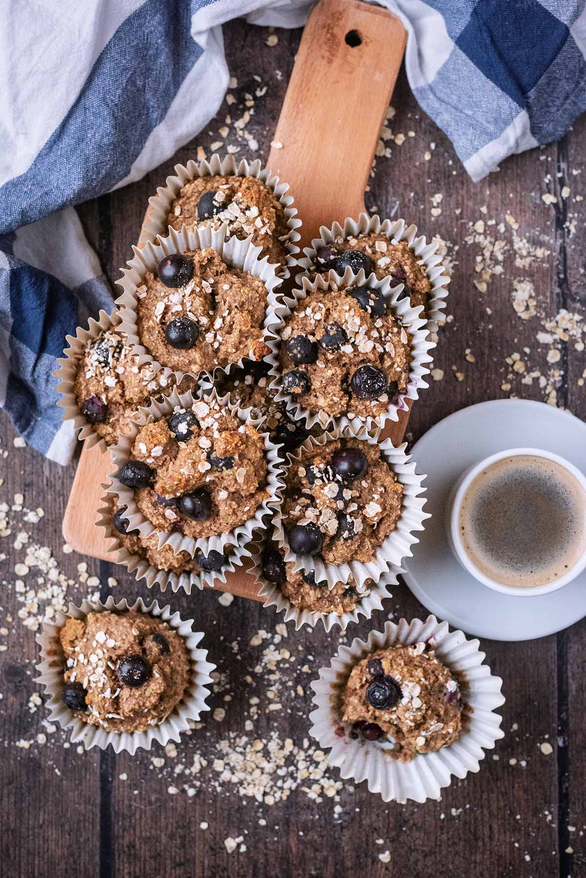 A pile of blueberry muffins on a wooden board next to a cup off black coffee.