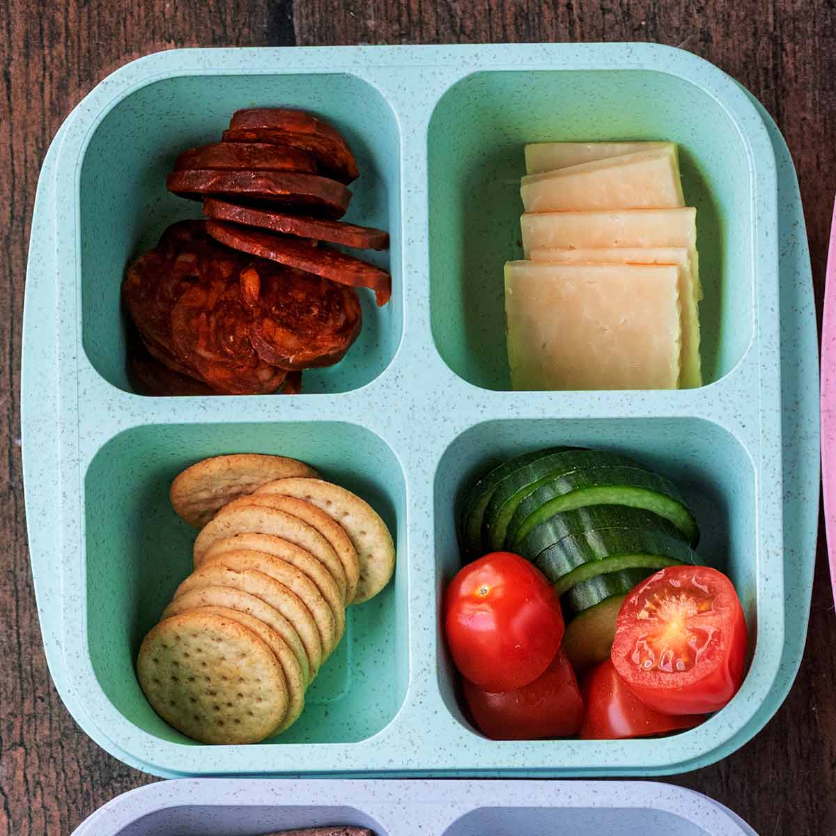 A four compartment snack box containing chorizo, cheese, crackers, tomatoes and cucumber.