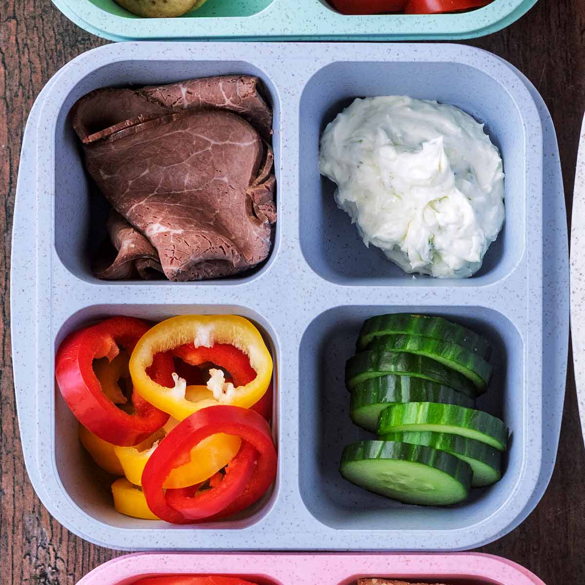 A four compartment snack box containing beef slices, tzatziki, pepper slices and cucumber slices.